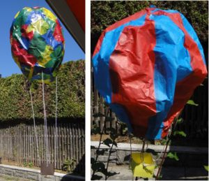 In diesem Bild sind zwei gebastelte Heißluftballons gezeigt. Links ist einer aus Transparentpapier gebastelt. ER ist sehr bunt und hängt in der Luft. Daneben ist ein rot-blauer Ballon aus Seidenpapier. Beiden haben einen Korb am Ende einer Schnur hängen.