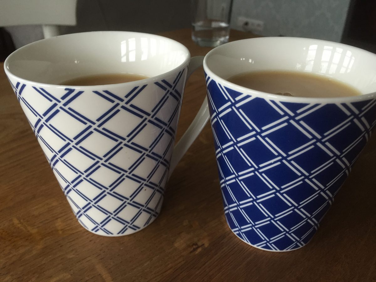 Zu sehen sind zwei Teetassen. Eine weiß mit blauem Karomuster, eine blau mit weißem Karomuster. Beide sind mit Schwarztee gefüllt