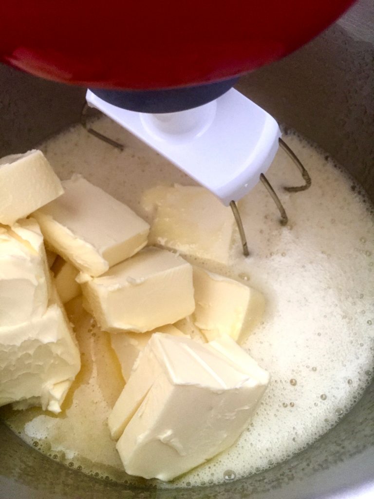 In einer Rührschüssel ist die schaumige Eimasse und die Butter zu sehen. Der Rührhakten steckt schon in der zu mixenden Masse.