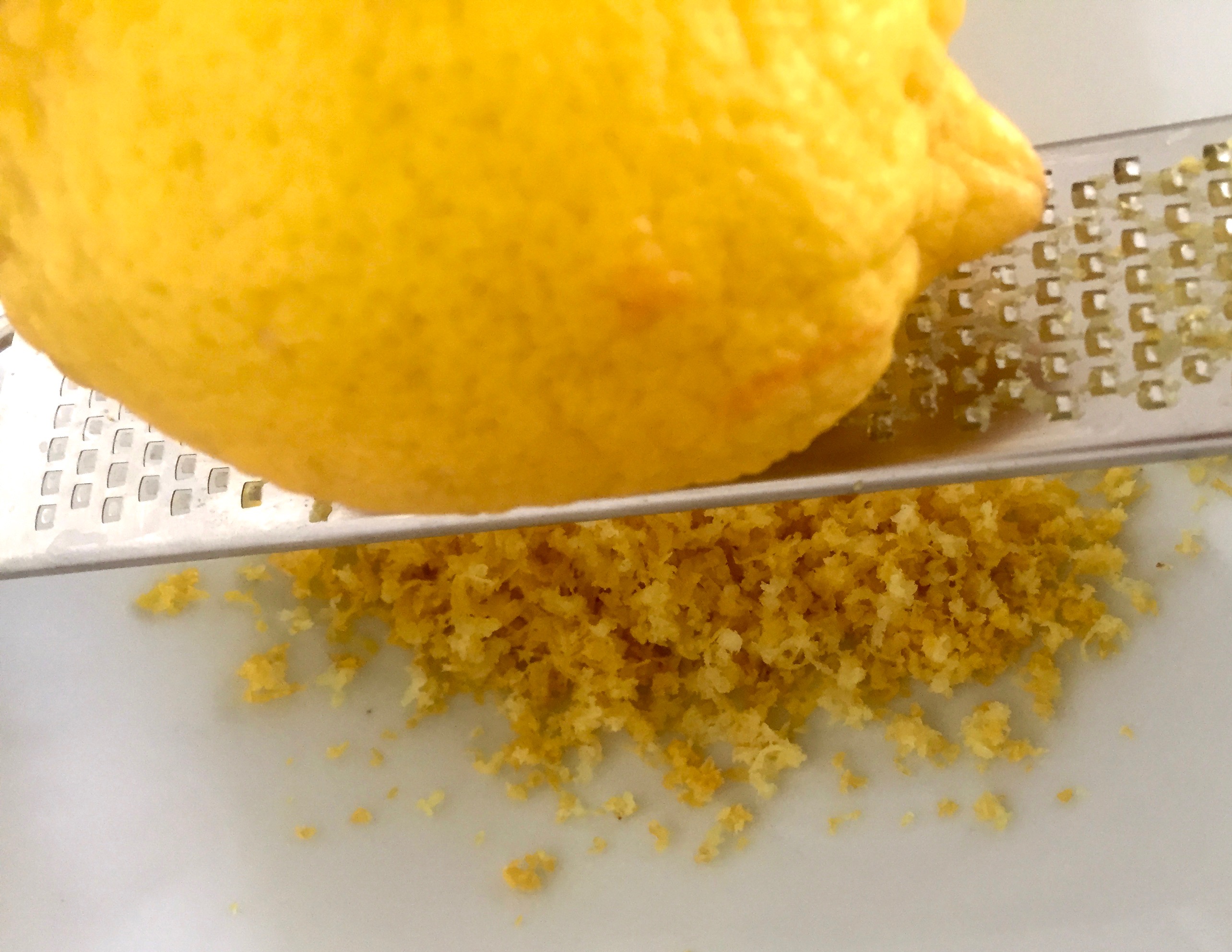 Die Schale einer Zitronen wird abgerieben. Dazu liegt eine Zitrone auf einer Reibe und darunter wird die Schale aufgefangen.