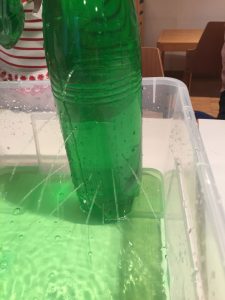 In einer mit grünem Wasser gefüllten Flasche spritzt Wasser aus kleinen Löchern in eine Auffangschale. Je höher das Loch in der Flasche ist, umso flacher strömt das Wasser nach unten.