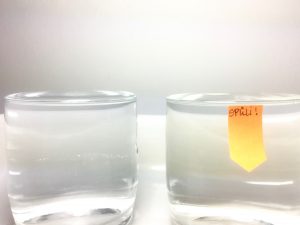 Auf dem Bild sind zwei Gläser zu sehen. Links ein Glas, das fast übervoll mit Wasser befüllt ist. Auf der rechten Seite ein Glas, das ebenfalls mit Wasser befüllt ist. In diesem ist Spülmittel enthalten und deshalb ist das Wasser nicht mehr so gespannt, wie im linken Wasserglas. Mit einem orange farbenen Schild ist auf das Spüli im Glas hingewiesen.