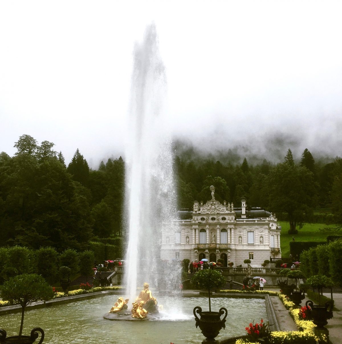 Blick auf das Schloss Linderhof. Im Vordergrund ist der große Teich mit riesiger Fontäne zu erkennen. Bunte Blumen schmücken den Schlossgarten.
