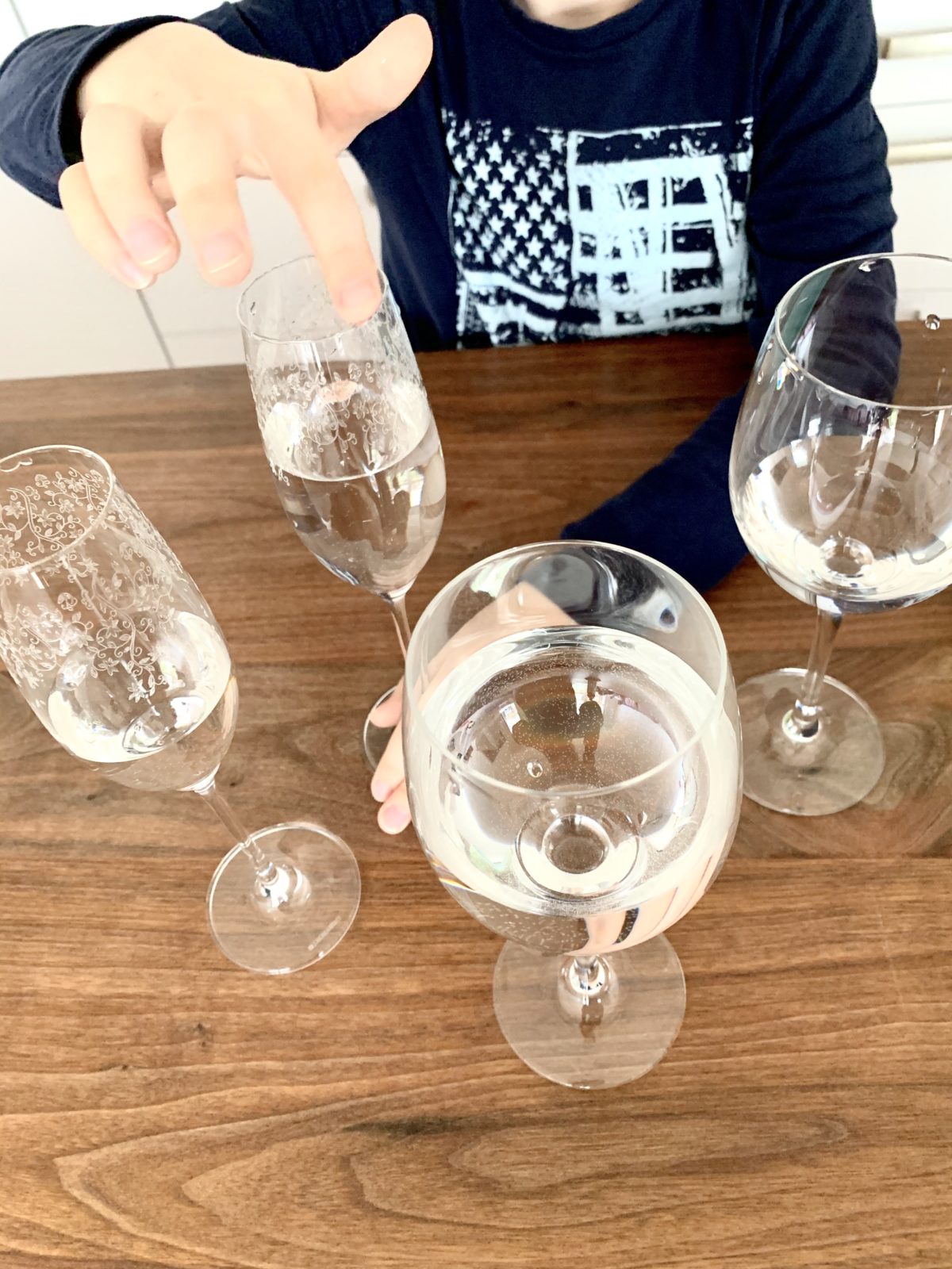 Mit Wasser gefüllte Weingläser und Sektgläser stehen auf einem Holztisch. Eines wird mit dem Finger am Glasrand umkreist, um einen Ton zu erzeugen.