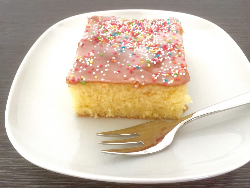 Ein Stück Zitronenkuchen mit Zuckerglasur und bunten Zuckerperlen liegt auf einem weißen Teller samt Kuchengabe.