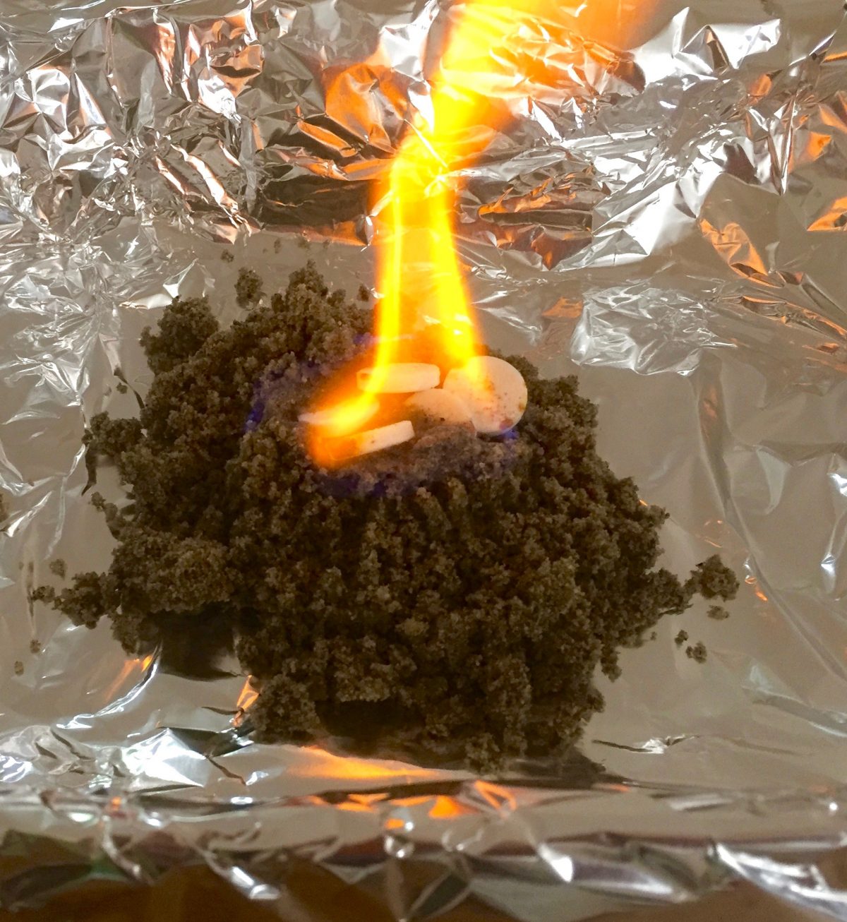Orange leuchtende Flamme über einem Sandhäufchen. Alufolie zum Schutz druntergelegt.