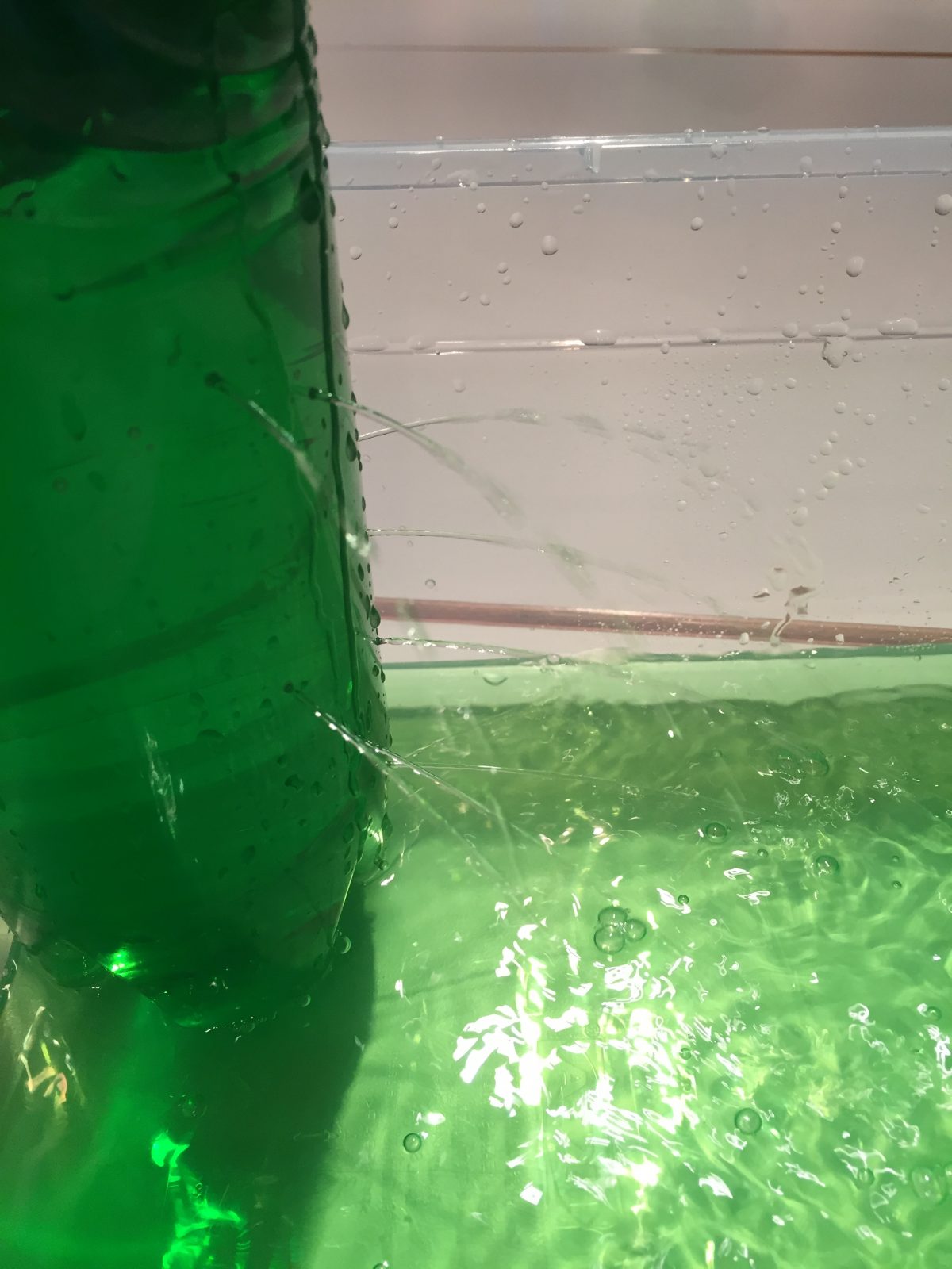 Aus einer Plastikflasche strömen Wasserstrahlen auf unterschiedlicher Höhe heraus. Das Wasser ist grün angefärbt und wird in einer Schale aufgefangen.