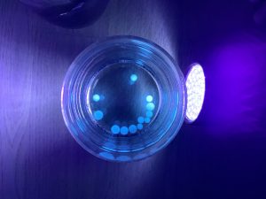 Wasserperlen liegen in Form eines Gesichtes in einem Glas und werden mit UV-Licht angestrahlt. Dadurch leuchten sie hellblau.